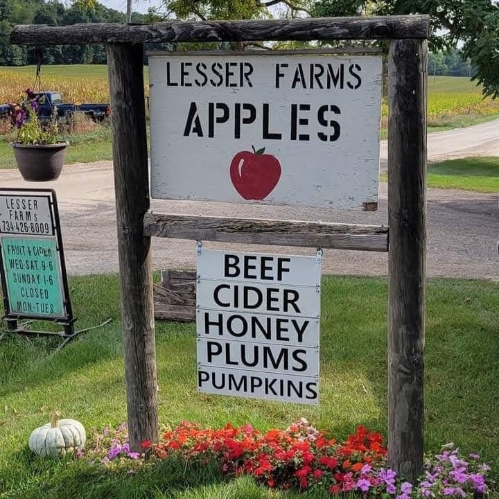 Lesser Farms sign displaying apples and additional products for sale, including beef, cider, honey, plums, and pumpkins, with operating hours listed.