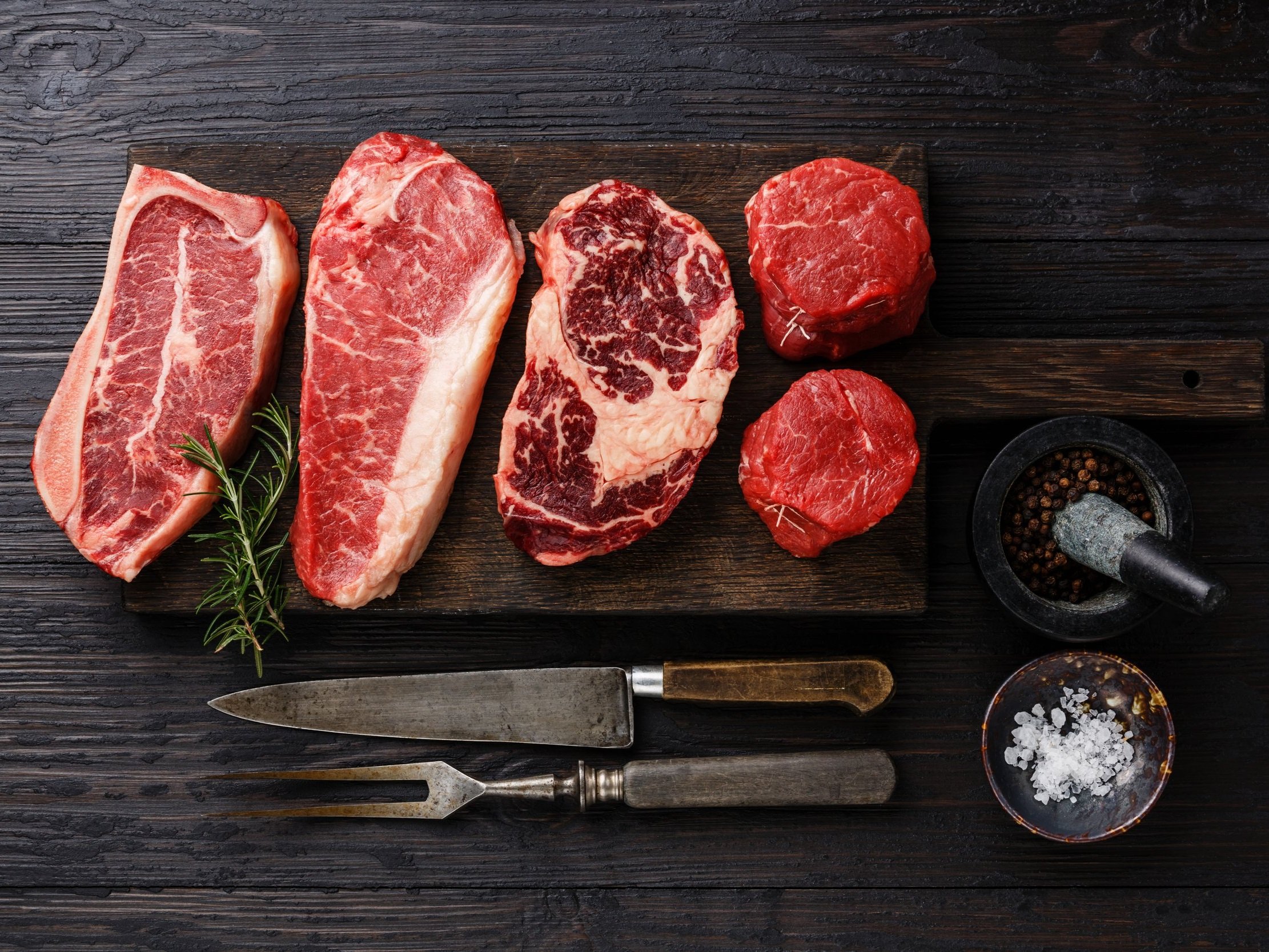 Selection of premium beef cuts including ribeye, sirloin, and tenderloin on a wooden cutting board, accompanied by fresh rosemary, a mortar and pestle with peppercorns, and a bowl of sea salt. Ideal for gourmet cooking and grilling.