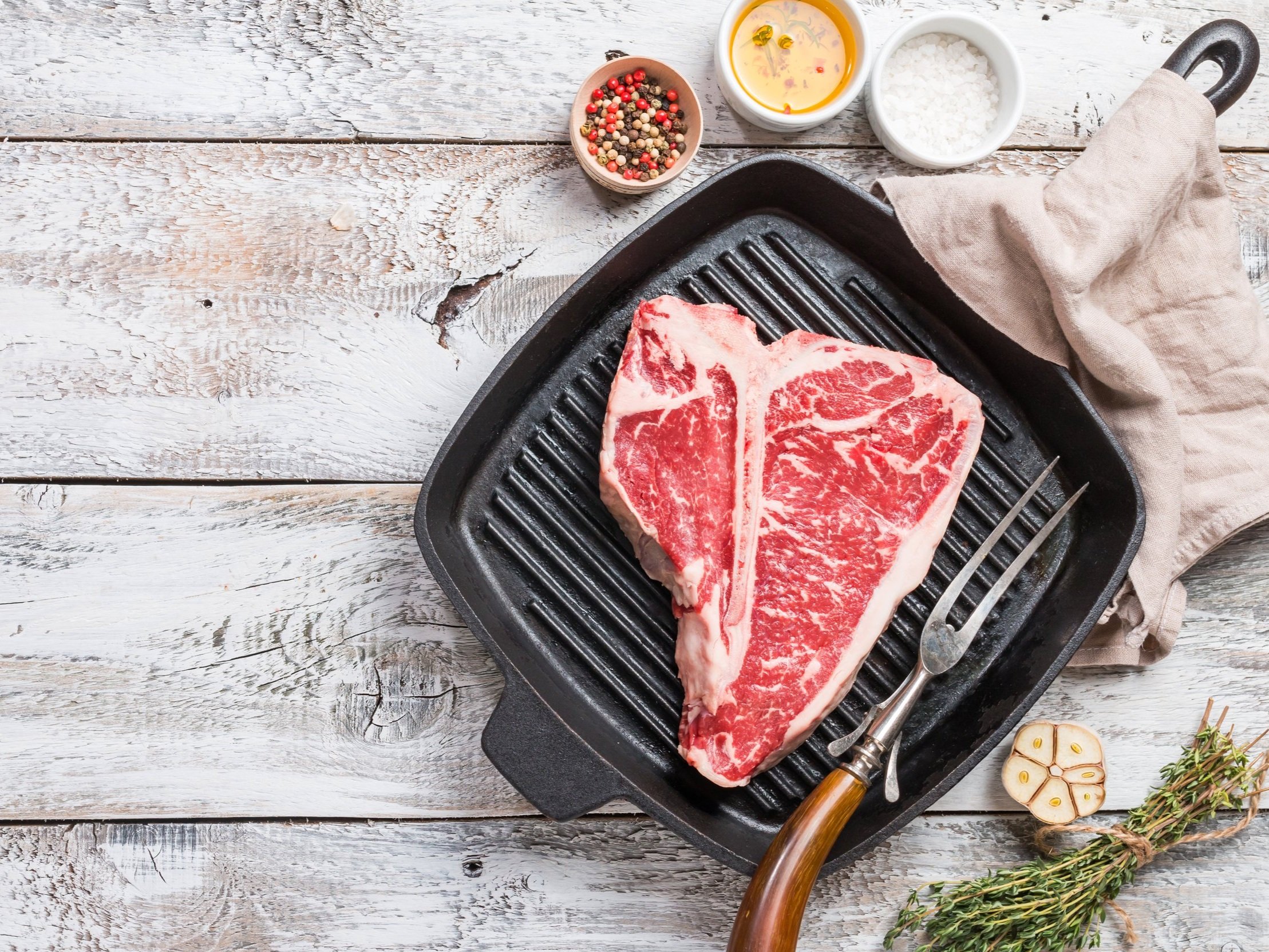 Juicy T-bone steak on a cast iron grill pan, surrounded by fresh herbs, garlic, and small bowls of olive oil, salt, and assorted peppercorns, showcasing a rustic kitchen setting.